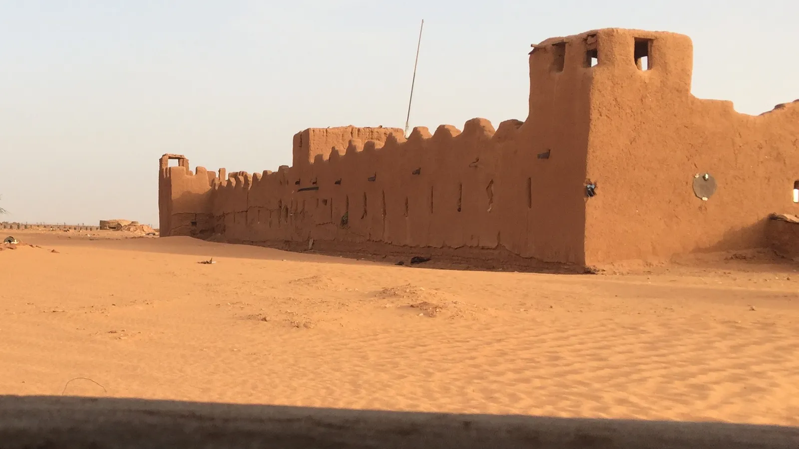 The mud-brick walls and tower of Fort Madama rising from orange sand dunes in northern Niger under a pale sky