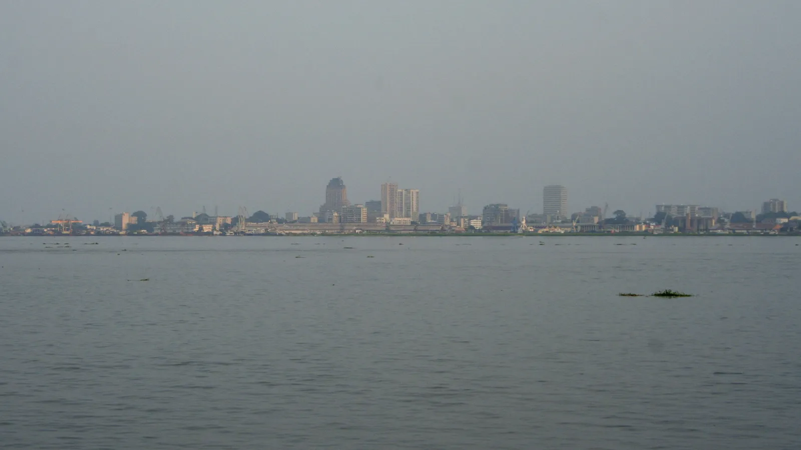 The Kinshasa skyline, low and grey on the far bank of the wide Congo River, seen from Brazzaville under a hazy sky